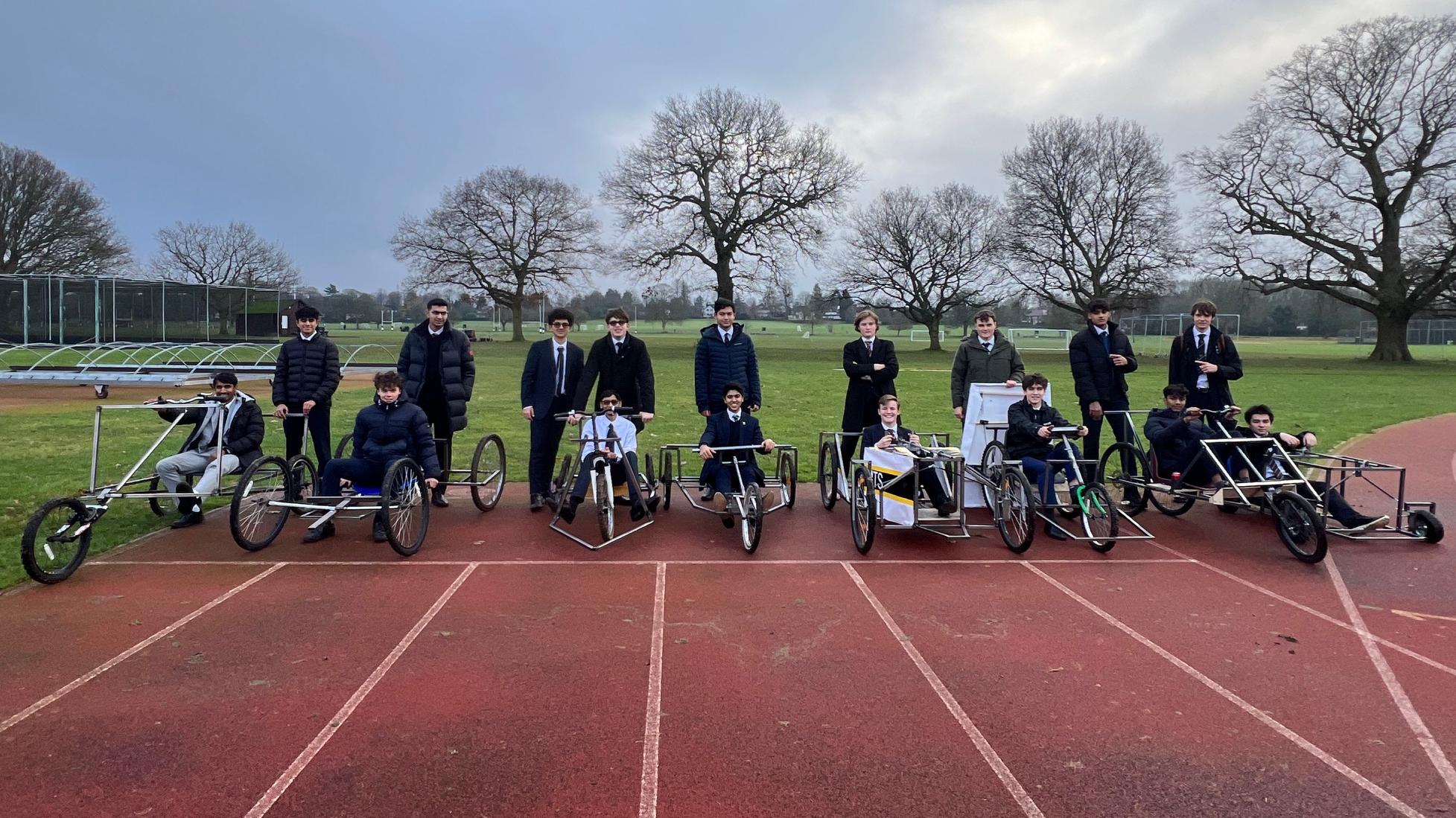 A group of students on a running track sat in and stood next to soap box racers they have made